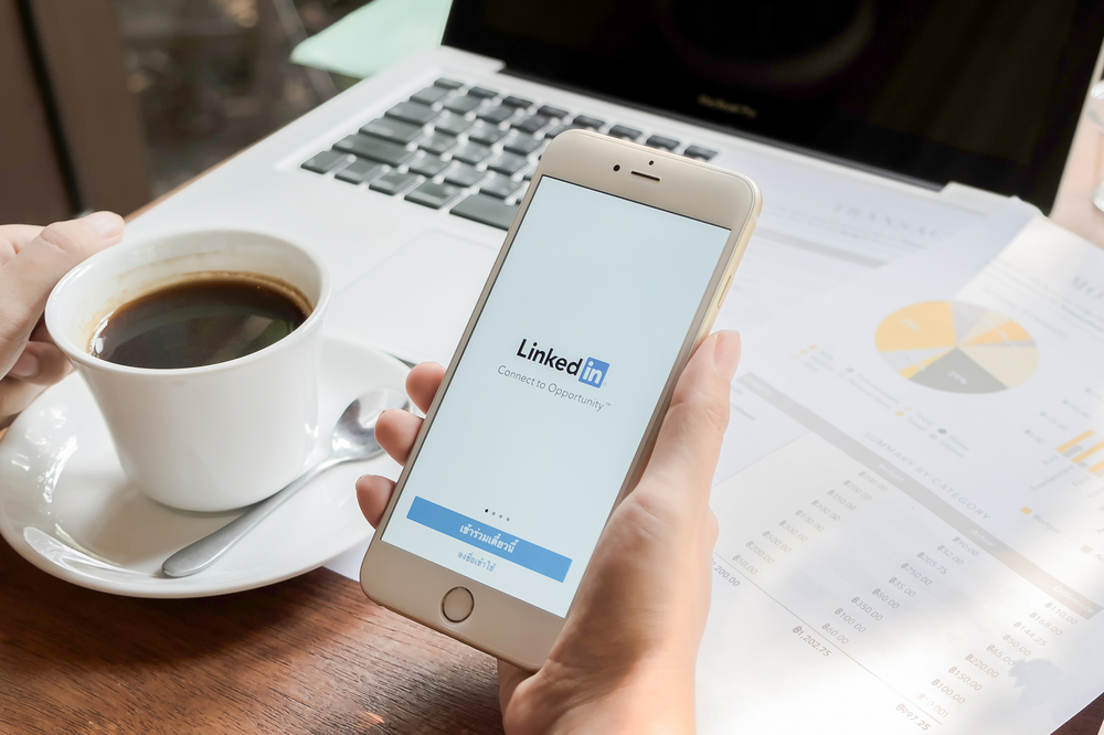 Persons hands at laptop holding a phone open to LinkedIn and cup of coffee